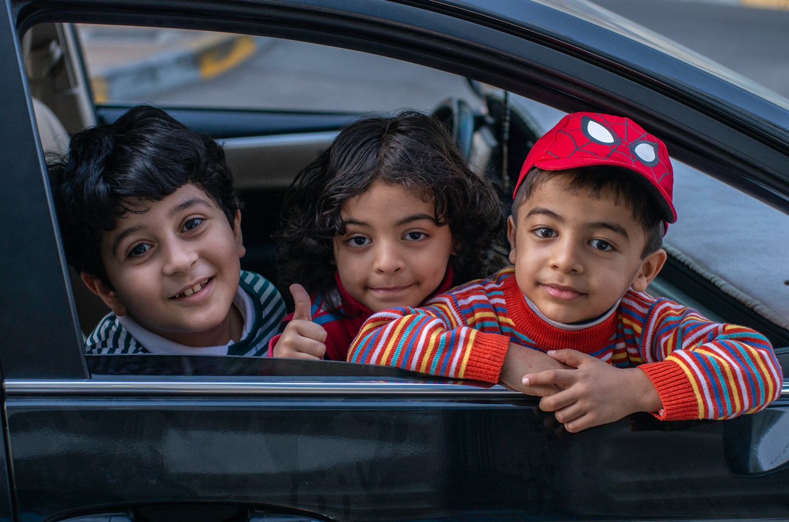 Three smiling children leaning out of a car window, one giving a thumbs up
